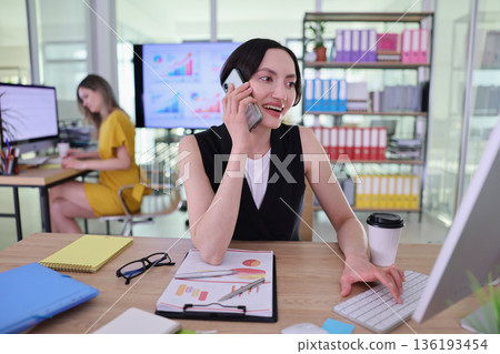 Young businesswoman engaged in a lively phone call while working in a modern office filled with vibrant charts and organized supplies 136193454