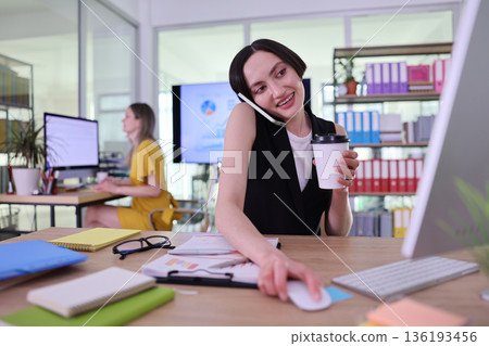 Busy office scene with a woman multitasking on calls while sipping coffee and working at her desk in a creative environment Busy office scene with a woman multitasking on calls while sipping coffee and working at her desk in a creative environment 136193456