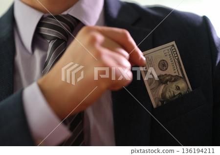 Businessman discreetly places a hundred-dollar bill in his suit pocket during a meeting in a modern office setting Businessman discreetly places a hundred-dollar bill in his suit pocket during a meeting in a modern office setting 136194211