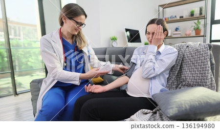 Healthcare professional checks blood pressure of pregnant woman in cozy living room during sunny day 136194980