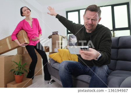 Emotional tension unfolds in a messy living room as a woman carries boxes while a man reacts to a phone call 136194987