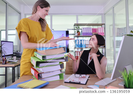 Two women engage in a dynamic discussion surrounded by colorful stacks of files in a modern office environment Two women engage in a dynamic discussion surrounded by colorful stacks of files in a modern office environment 136195059
