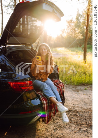 Young curly woman in stylish clothes in the trunk of a car in the forest and uses the phone. 136195546