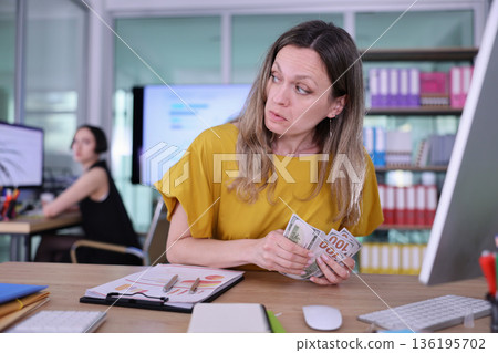 Uneasy office scene reveals tension as a woman discreetly counts cash behind a computer while a coworker focuses on her tasks nearby 136195702