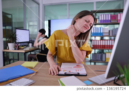 Office worker struggles with neck pain while analyzing reports during a busy workday in a modern office space 136195724