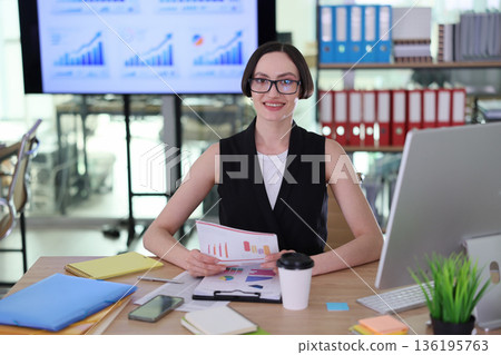 Confident professional reviewing charts and graphs in a modern office setting during a busy workday 136195763