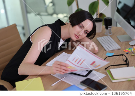 Focused woman reviewing charts and documents at a modern office desk during a busy workday Focused woman reviewing charts and documents at a modern office desk during a busy workday 136195764