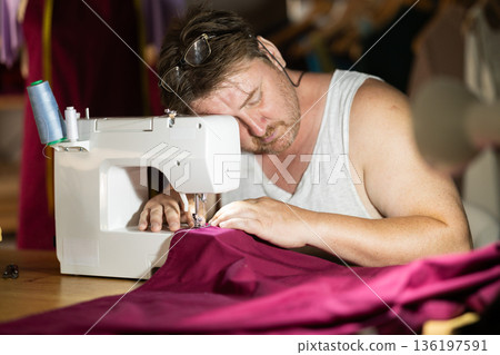Late in evening, tired man tailor dozed off next to sewing machine. 136197591