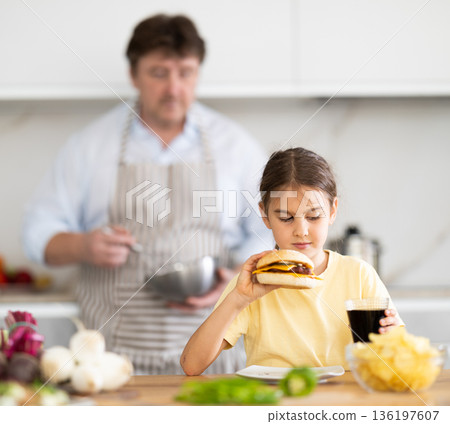 Little daughter eats meat burger and drinks carbonated drink in the kitchen. In background, father prepares healthy vegetable salad 136197607