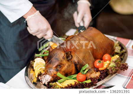 Roast pig with vegetables, gloved chef's hands cut with a knife Roast pig with vegetables, gloved chef's hands cut with a knife 136201391