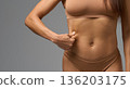 Close-up of woman in beige underwear pinching skin on side of stomach in studio light. 136203175