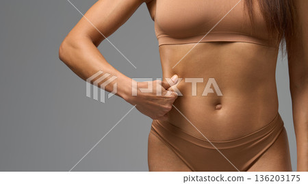 Close-up of woman in beige underwear pinching skin on side of stomach in studio light. 136203175