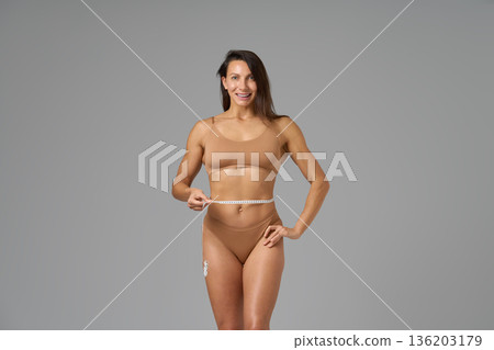 Full-length portrait of fit woman measuring waist with tape showing confidence and body positivity 136203179