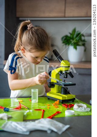 Young girl carefully placing chemical solution on microscope slide with pipette at home laboratory table. Preparation step for microscopic observation and hands on STEM chemistry experiment. 136204113