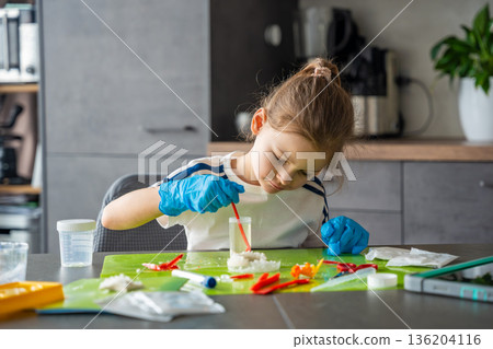 Young girl stirring chemical solution with measuring spoon in cup during crystal growing experiment at home. Chemistry practice and early STEM education through practical laboratory activity. Young girl stirring chemical solution with measuring spoon in cup during crystal growing experiment at home. Chemistry practice and early STEM education through practical laboratory activity. 136204116