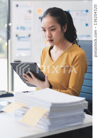 Asian woman works on laptop at office desk. She smiles happily while typing on keyboard near window with blinds. Professional woman works in business environment. Asian woman works on laptop at office desk. She smiles happily while typing on keyboard near window with blinds. Professional woman works in business environment. 136204320
