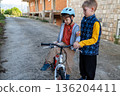 Boy helping his friend to sit on a bicycle outdoors, support and friendship, kids learning to ride a bike, child safety. 136204411