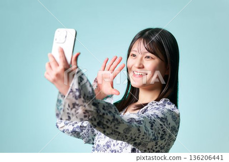 A woman in her 20s waving her hand at her smartphone screen and smiling brightly 136206441