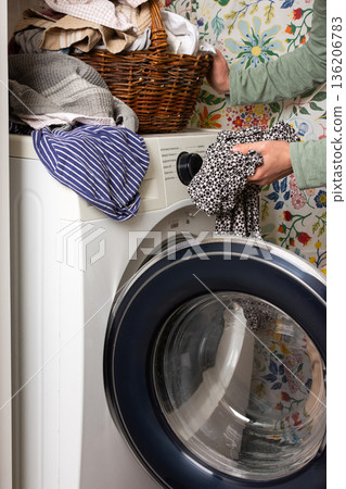 A woman sorts and loads laundry into a washing machine. The vibrant floral wall adds warmth to the cozy room. A basket of clothes sits nearby, ready to be washed A woman sorts and loads laundry into a washing machine. The vibrant floral wall adds warmth to the cozy room. A basket of clothes sits nearby, ready to be washed 136206783