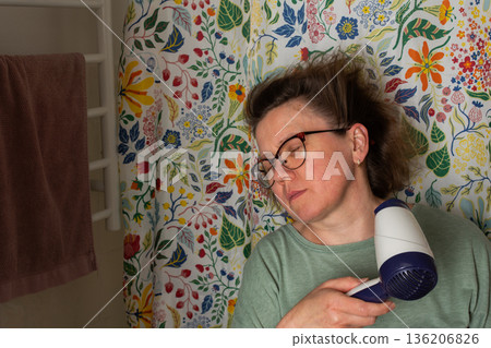 A woman prepares for the day in her bathroom. She is using a hair dryer to style her hair while standing in front of a vibrant shower curtain. Morning light fills the room A woman prepares for the day in her bathroom. She is using a hair dryer to style her hair while standing in front of a vibrant shower curtain. Morning light fills the room 136206826