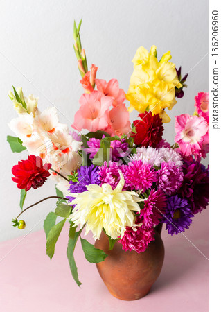 A clay vase holds a mix of flowers including glads and daisies. The vase is placed on a pink table. The room has bright light coming from outside 136206960