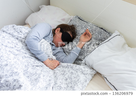 A 50-year-old woman is sound asleep in her bed, wearing a sleep mask. She is surrounded by pillows and blankets, creating a tranquil atmosphere. Sunlight gently filters into the room 136206979