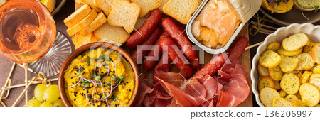 A vibrant charcuterie board showcases a variety of cheese, sausages, hummus, nuts, and sandwiches. Friends gather at a sunny outdoor picnic, enjoying delicious food together, banner A vibrant charcuterie board showcases a variety of cheese, sausages, hummus, nuts, and sandwiches. Friends gather at a sunny outdoor picnic, enjoying delicious food together, banner 136206997