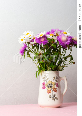A colorful bouquet of daisies and purple flowers arranged in a charming pitcher rests on a pink tablecloth, bringing joy to the soft, simple setting A colorful bouquet of daisies and purple flowers arranged in a charming pitcher rests on a pink tablecloth, bringing joy to the soft, simple setting 136207056