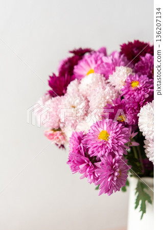 Bright flowers are arranged in a white vase with a floral pattern. The vase is placed on a light surface, showcasing the colorful blooms during the day in a simple space 136207134