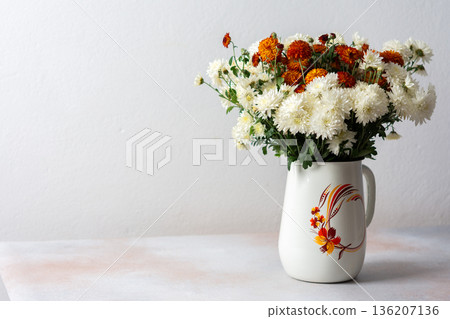 A white pitcher filled with orange and white flowers sits on a light surface. This setup can be used for home decoration or floral arrangement inspiration 136207136