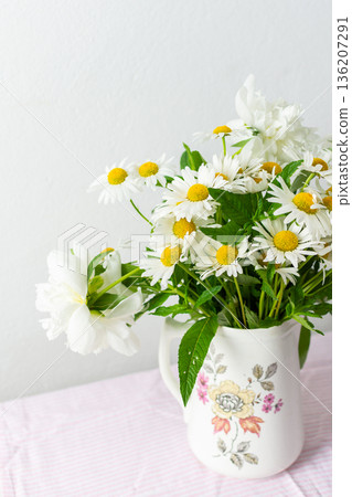 Fresh daisies are arranged in a decorative vase, brightening up a light room. The soft pink tablecloth enhances the cheerful atmosphere, showcasing spring flowers Fresh daisies are arranged in a decorative vase, brightening up a light room. The soft pink tablecloth enhances the cheerful atmosphere, showcasing spring flowers 136207291