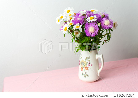 A colorful bouquet of daisies and purple flowers arranged in a charming pitcher rests on a pink tablecloth, bringing joy to the soft, simple setting A colorful bouquet of daisies and purple flowers arranged in a charming pitcher rests on a pink tablecloth, bringing joy to the soft, simple setting 136207294