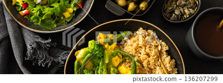 Healthy breakfast consists of a quinoa bowl with boiled eggs, green beans, and broccoli placed on a dark surface next to a cup of coffee and a woven cloth, banner 136207299