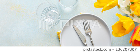 A fresh spring table setting features a plain plate, silver utensils, and a bright yellow napkin. Colorful flowers in bloom add a lively touch, creating a welcoming atmosphere for meals, banner 136207307