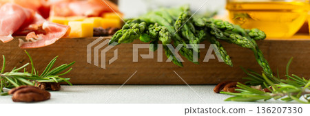 A vibrant charcuterie board is set for a gathering, showcasing fresh asparagus, assorted cheeses, cured meats, and nuts. This setup invites guests to enjoy tasty bites together, banner 136207330