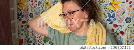 A 50-year-old woman starts her morning routine in the bathroom. She looks at herself in the mirror while holding a towel over her shoulder. The colorful background adds brightness, banner 136207331