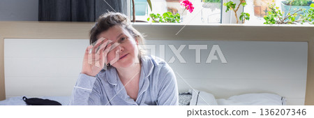 A tired 50-year-old woman sits on her bed, rubbing her eyes as she adjusts to the early morning light. Her cozy bedroom features plants and flowers visible outside the window, banner A tired 50-year-old woman sits on her bed, rubbing her eyes as she adjusts to the early morning light. Her cozy bedroom features plants and flowers visible outside the window, banner 136207346