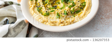 A bowl of creamy hummus topped with olive oil and herbs sits beside fresh bread and a bowl of olives. The light setting showcases a simple, inviting meal, banner 136207353