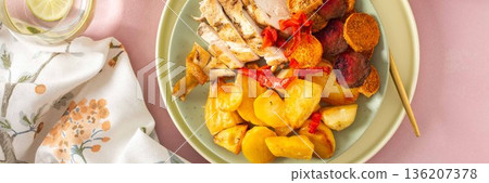 Baked potato is served with chicken breast and roasted carrots, peppers, and beets on a plate. A light meal is arranged on a simple table setting, banner 136207378
