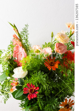 A bouquet of roses, gladioli, zinnias, and eustomas stands in water. Carrots with green tops are part of the arrangement on a plain table against a light background 136207385