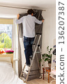 A man is using a ladder to install curtains on a rod in a well-lit room. He holds gray curtains in one hand while adjusting the rod above a window. Plants and flowers are seen outside 136207387