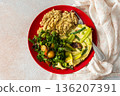 Grains, cooked greens, and vegetables are arranged on a red plate. A bowl of mixed salad with tomatoes is placed nearby on a light surface 136207391