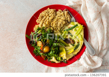 Grains, cooked greens, and vegetables are arranged on a red plate. A bowl of mixed salad with tomatoes is placed nearby on a light surface 136207391