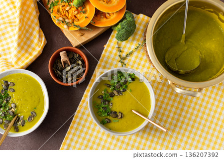 A table displays bowls of pumpkin soup with broccoli topped with seeds and herbs. Nearby, fresh ingredients and a pot hold more soup, creating a healthy meal setting A table displays bowls of pumpkin soup with broccoli topped with seeds and herbs. Nearby, fresh ingredients and a pot hold more soup, creating a healthy meal setting 136207392