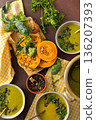 A table displays bowls of pumpkin soup with broccoli topped with seeds and herbs. Nearby, fresh ingredients and a pot hold more soup, creating a healthy meal setting 136207393