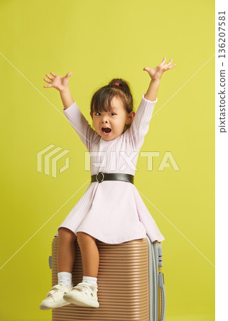Little joyful Asian girl sits on suitcase and cheerfully spreads her arms to up, expressing happiness from a long-awaited vacation and trip to the sea 136207581