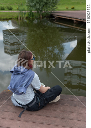Person sitting on wooden boardwalk by pond with reflection of modern building and green park surroundings 136208511