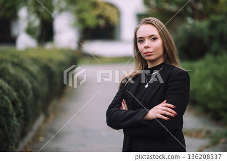 Confident Young Woman Standing with Crossed Arms in Park Wearing Black Jacket Natural Light Shallow Depth of Field and Soft Green Background 136208537