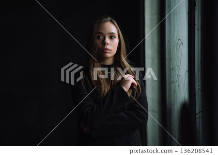 Moody Young Woman Portrait Standing Near Window Indoors with Natural Side Light and Dark Minimal Background Creating Cinematic Atmosphere Moody Young Woman Portrait Standing Near Window Indoors with Natural Side Light and Dark Minimal Background Creating Cinematic Atmosphere 136208541