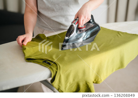 Woman ironing a green t-shirt clothing at home. Daily chores. Laundry day. Neutral background. Home appliances 136208849